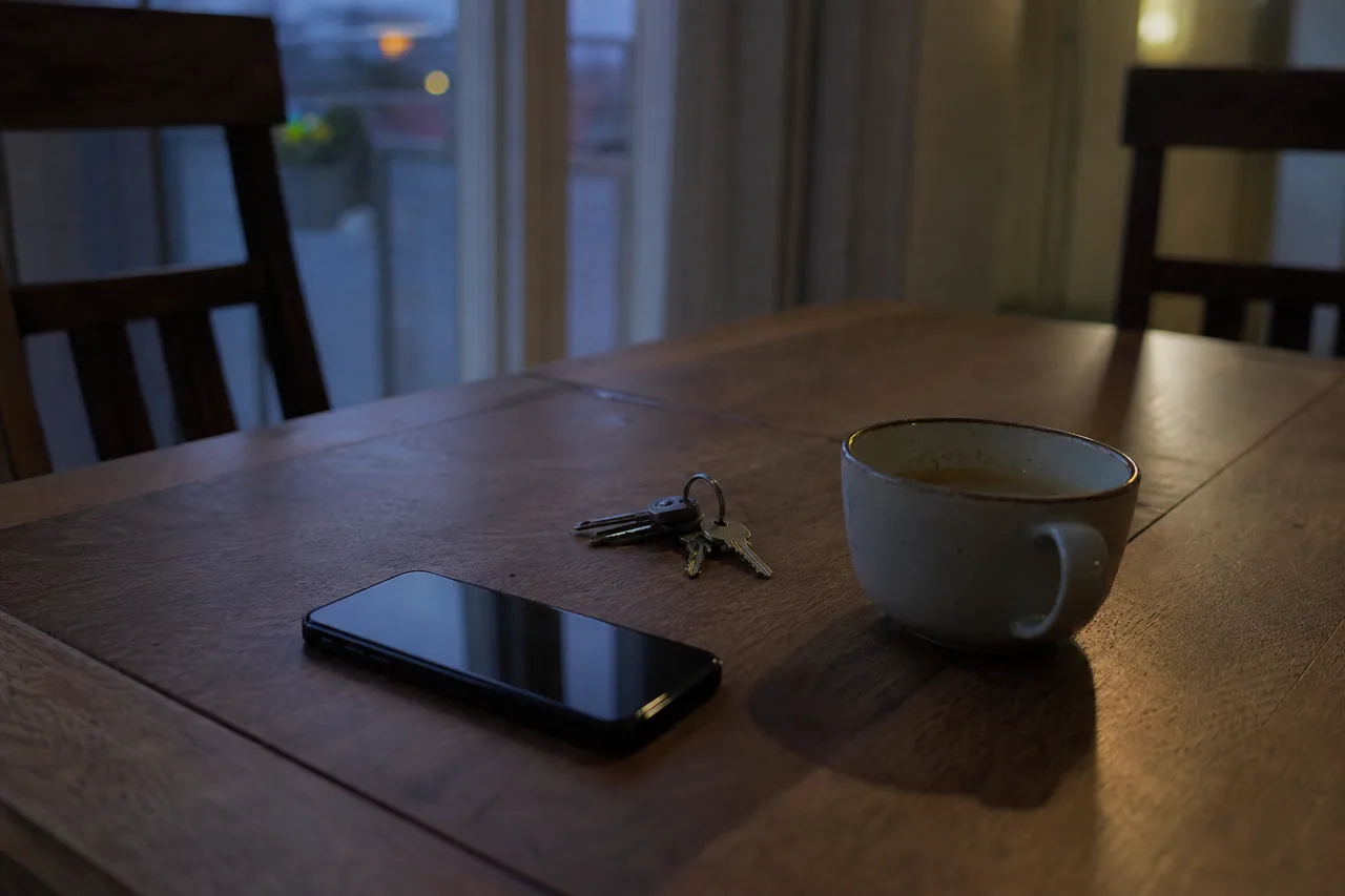 Telefon bimbit tertiarap di atas meja kayu bersebelahan kunci rumah dan cawan teh, cahaya petang