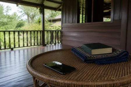 Beranda rumah kampung pada waktu pagi: telefon pintar, sejadah dan buku tertutup di atas meja rotan