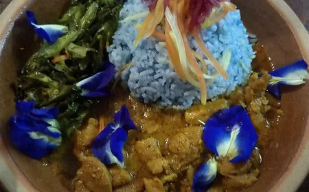 Semangkuk nasi kerabu biru bersama ulam, bunga telang, kari dan udang di atas meja kayu